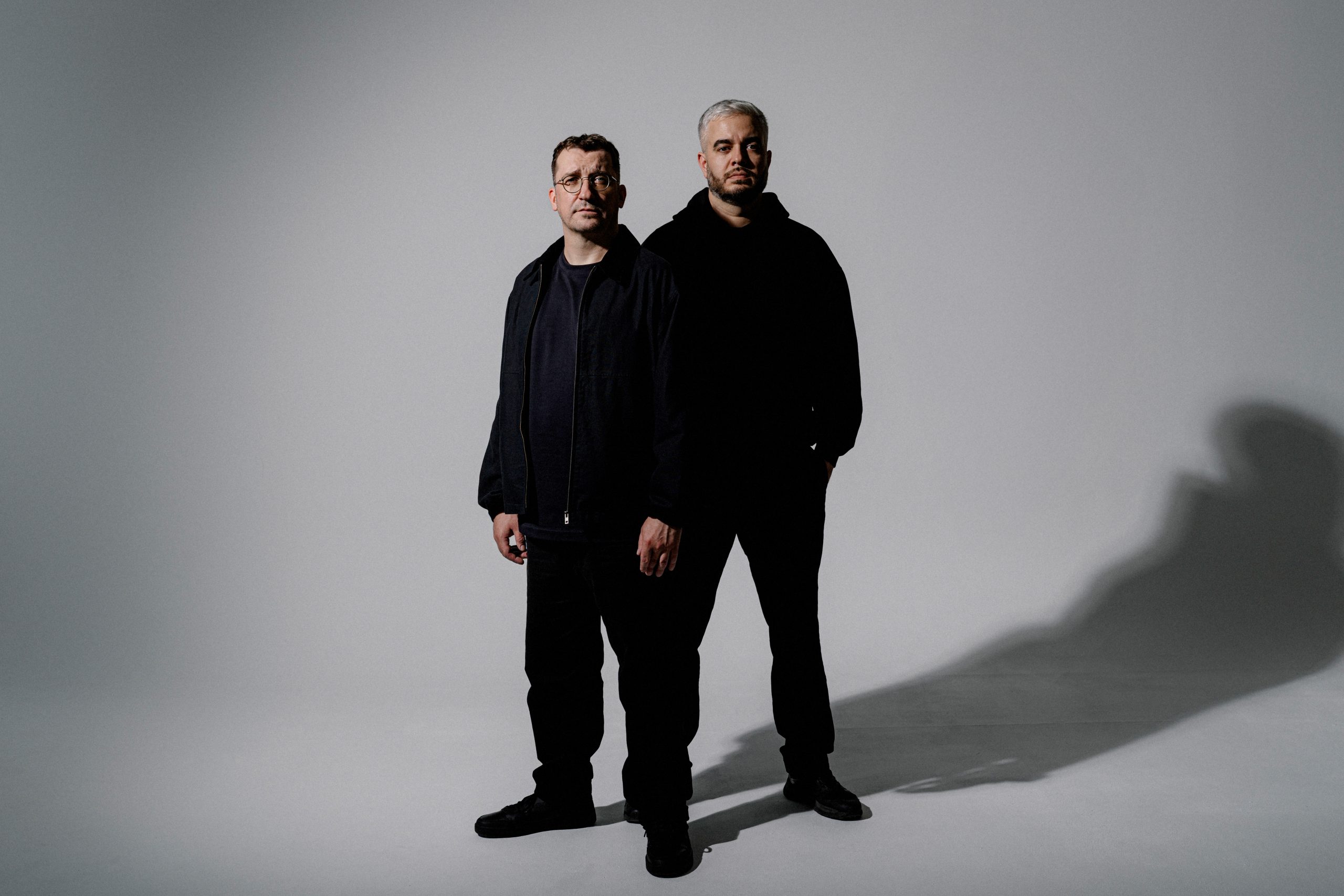 Two men stand side by side in a plain gray studio, wearing dark clothing and boots.