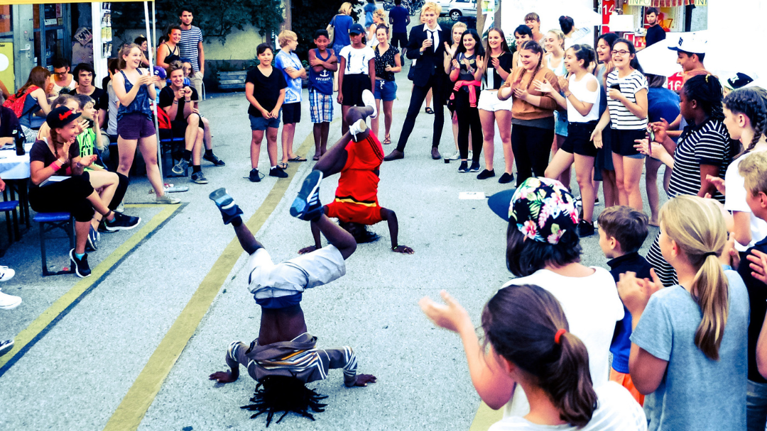 Crowd of onlookers forms a circle around three breakdancers performing headstands and freezes in a street performance.