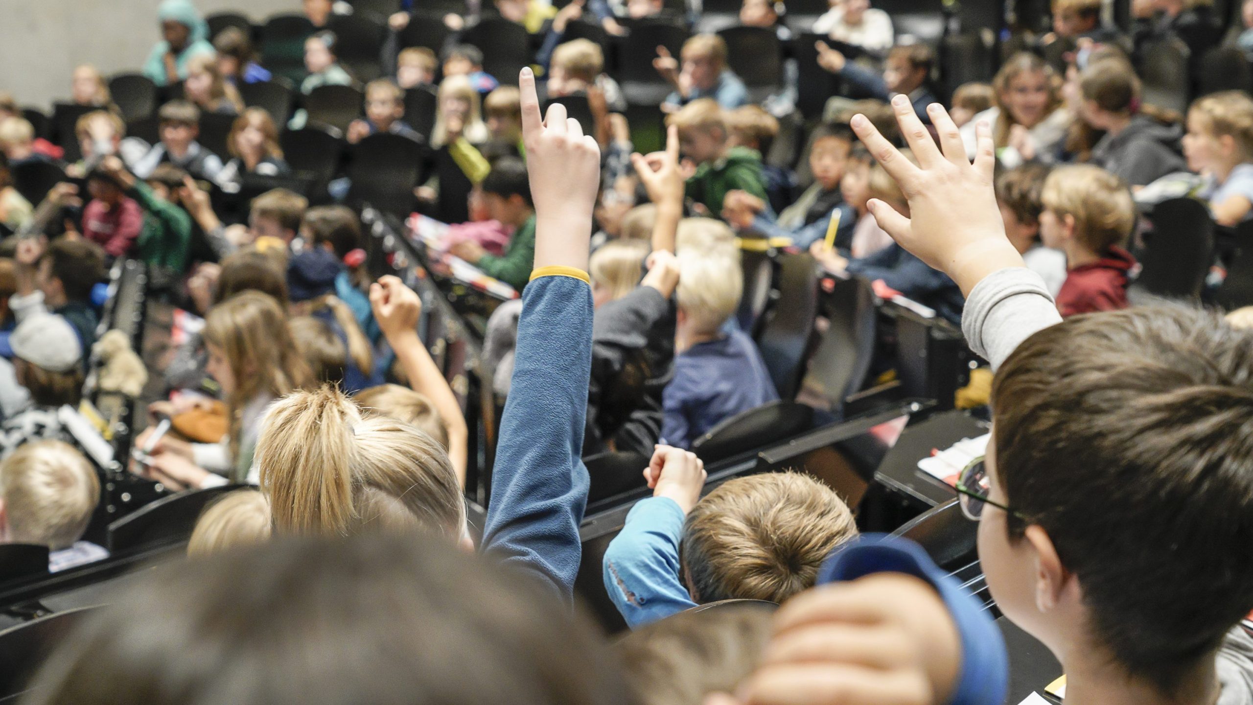 Students raise their hands in a crowded classroom or lecture hall, many seated with notebooks and pens nearby.