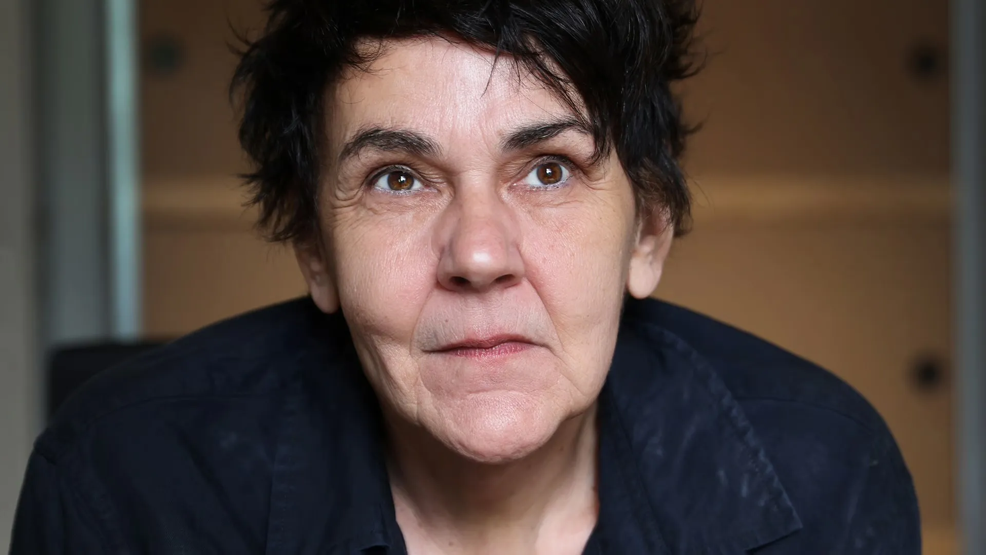 christiane Rösinger - IN München - Das Stadtmagazin Close-up portrait of a woman with short dark hair, looking upward with a thoughtful expression, wearing a dark shirt.