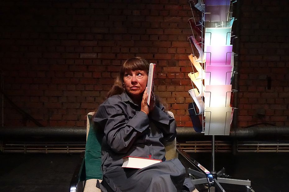 Woman in a gray coat sits in a studio chair, holding a notebook to her ear beside a tall rainbow-colored file rack against a brick wall.