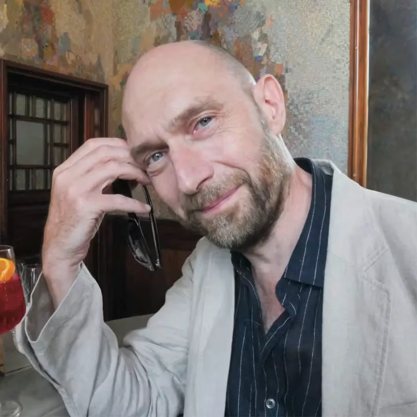 eric pfeil - IN München - Das Stadtmagazin Man with a shaved head and light beard sits at a table, holding sunglasses near his temple and smiling at the camera, with a drink on the table nearby.