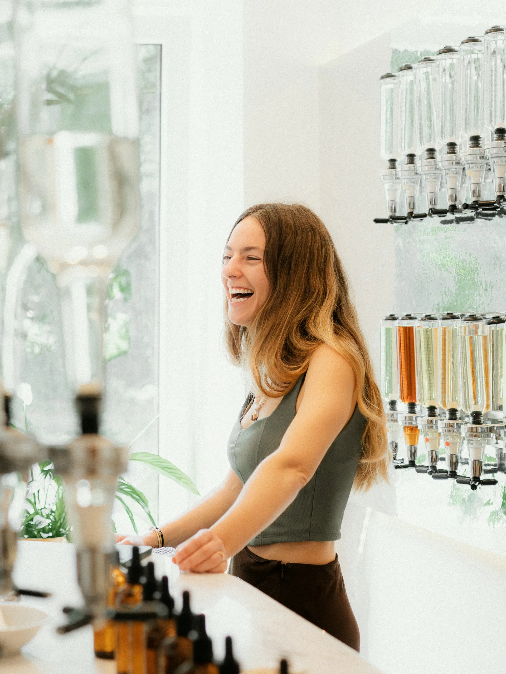 Woman with long hair laughing at a counter, with a row of glass beverage dispensers behind her.