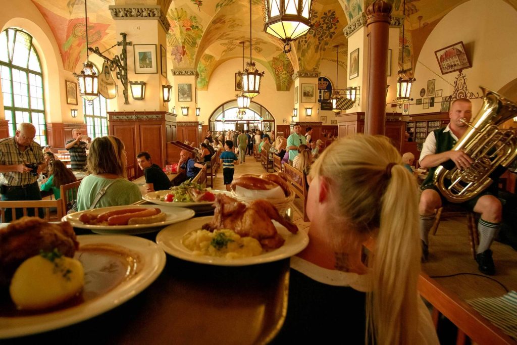 Busy Bavarian-style restaurant interior with long wooden benches, diners, and a tuba player on the right; plates of food in the foreground (from the diners' table).