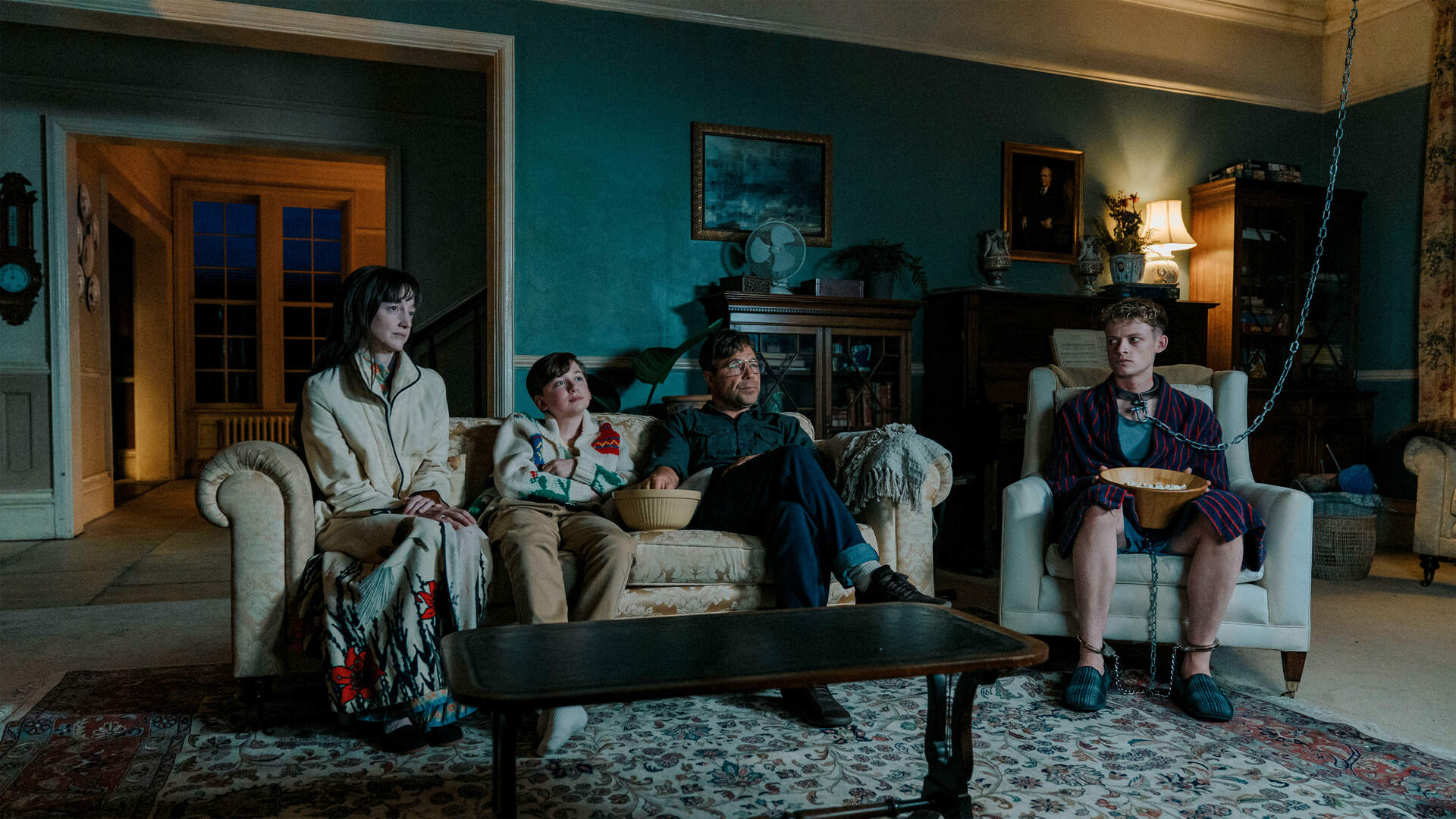 Dim living room with teal walls; four people sit on sofas, including a young man chained to a chair with a bowl nearby.