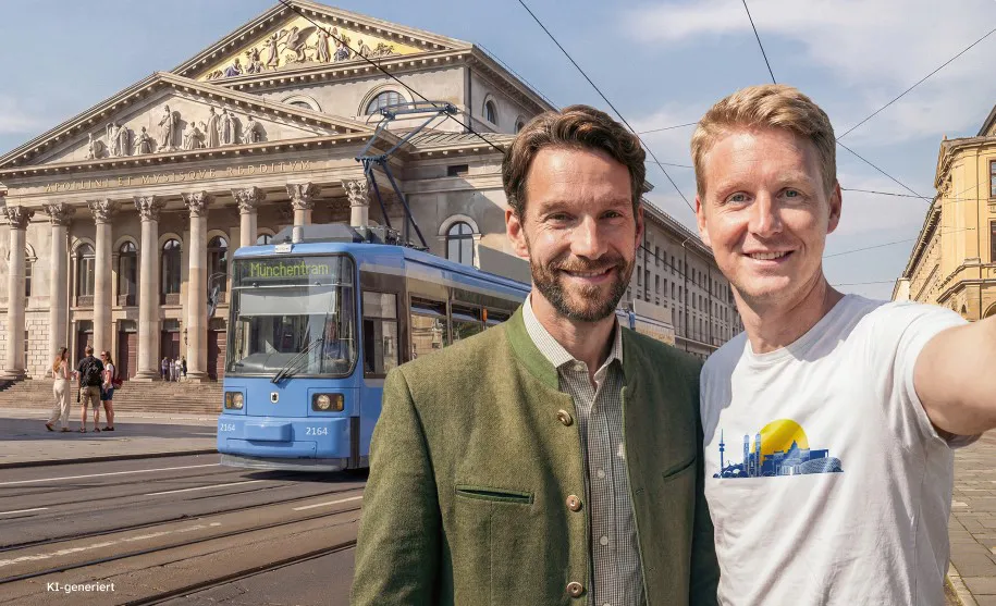 Two men take a selfie in front of a blue Munich tram with a grand historic building behind them.