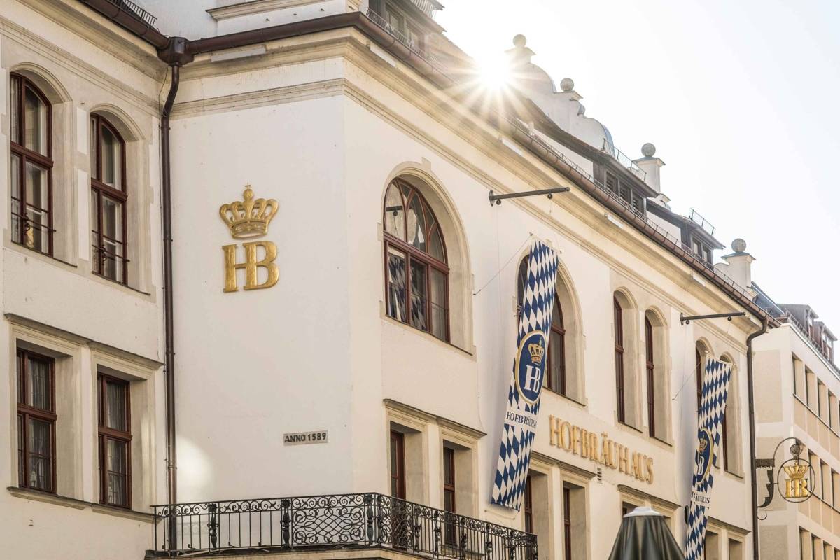 Exterior of Hofbräuhaus with HB crest and blue-and-white Bavarian flags on the white façade, sun glare above the roof.