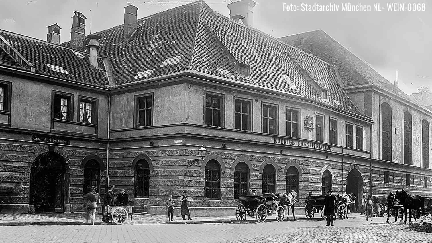 Historic Munich street scene: a large stone building with arched ground-floor windows and a tiled roof, with horse-drawn carriages and pedestrians on a cobbled street.