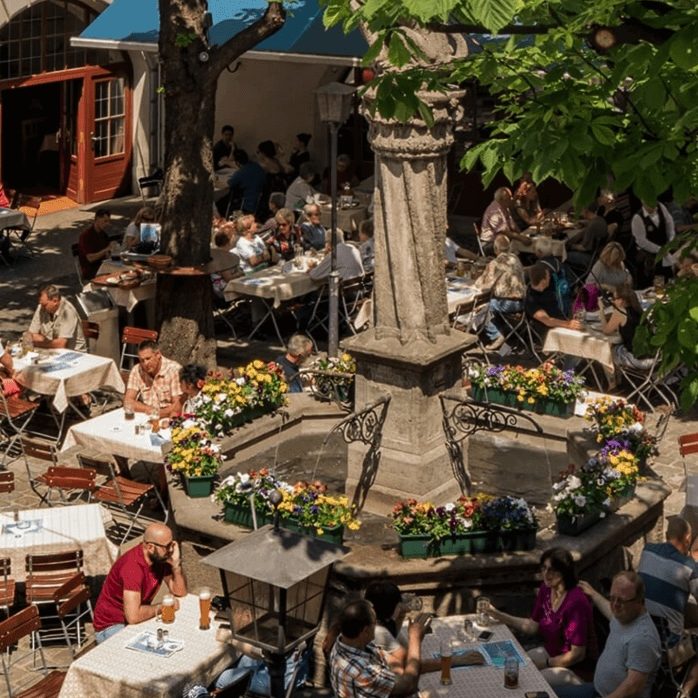 Outdoor cafe around a stone fountain, people dining at tables in a sunny plaza with flowers and trees nearby.
