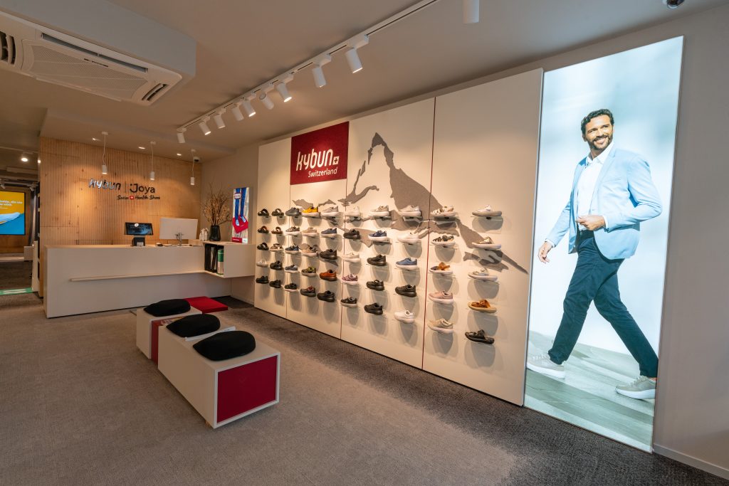 kJ-Shop_Muenchen_kybun-Wand - IN München - Das Stadtmagazin Shoe store interior with wall shelves displaying sneakers and a large poster of a smiling man in a blue suit on the right side.