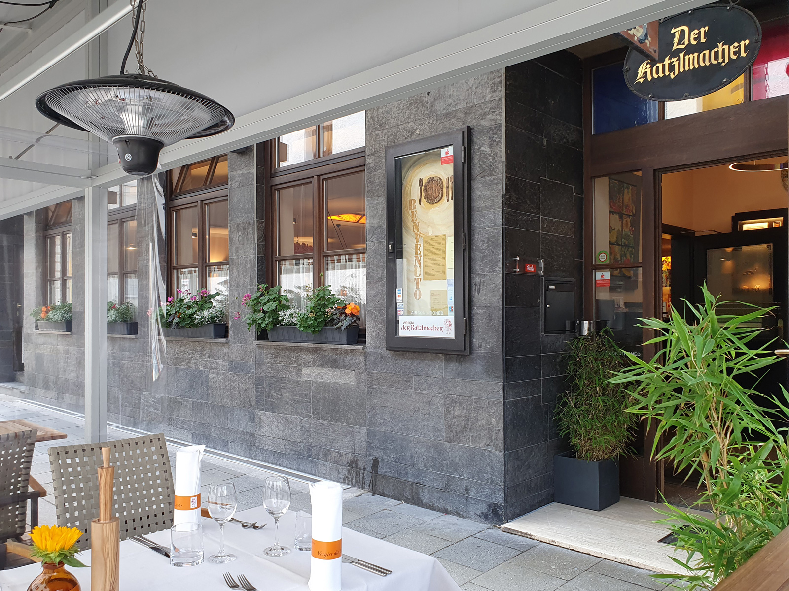 Outdoor restaurant terrace with white-tablecloth tables, glassware, and napkins, along a stone wall with flower boxes and a 'Der Katzlmacher' sign.