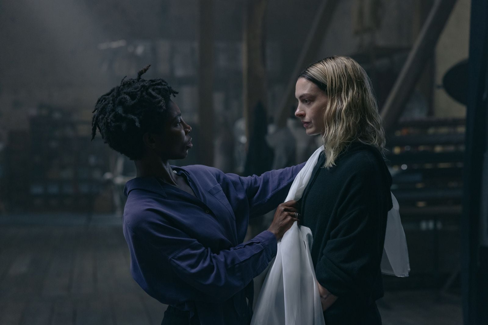 Two women in a dim workshop; one adjusts a white fabric draped over the other as if fitting a garment.