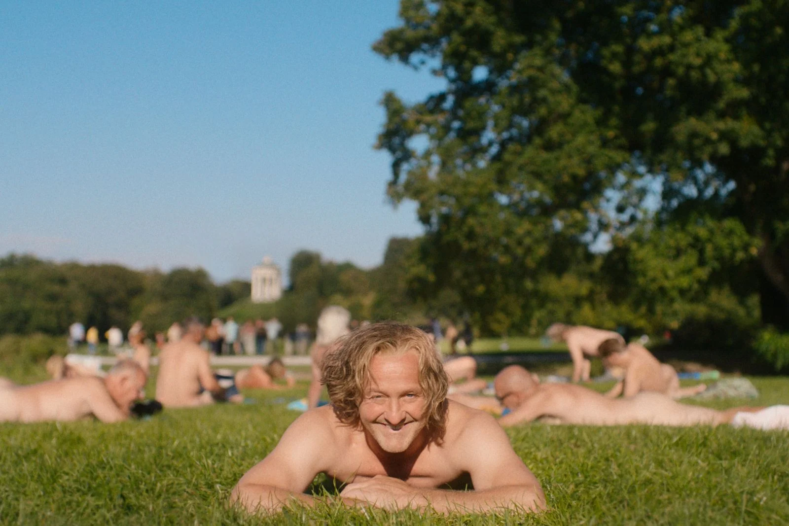 Smiling naked man lying on grass in a park, with other sunbathers visible in the background.