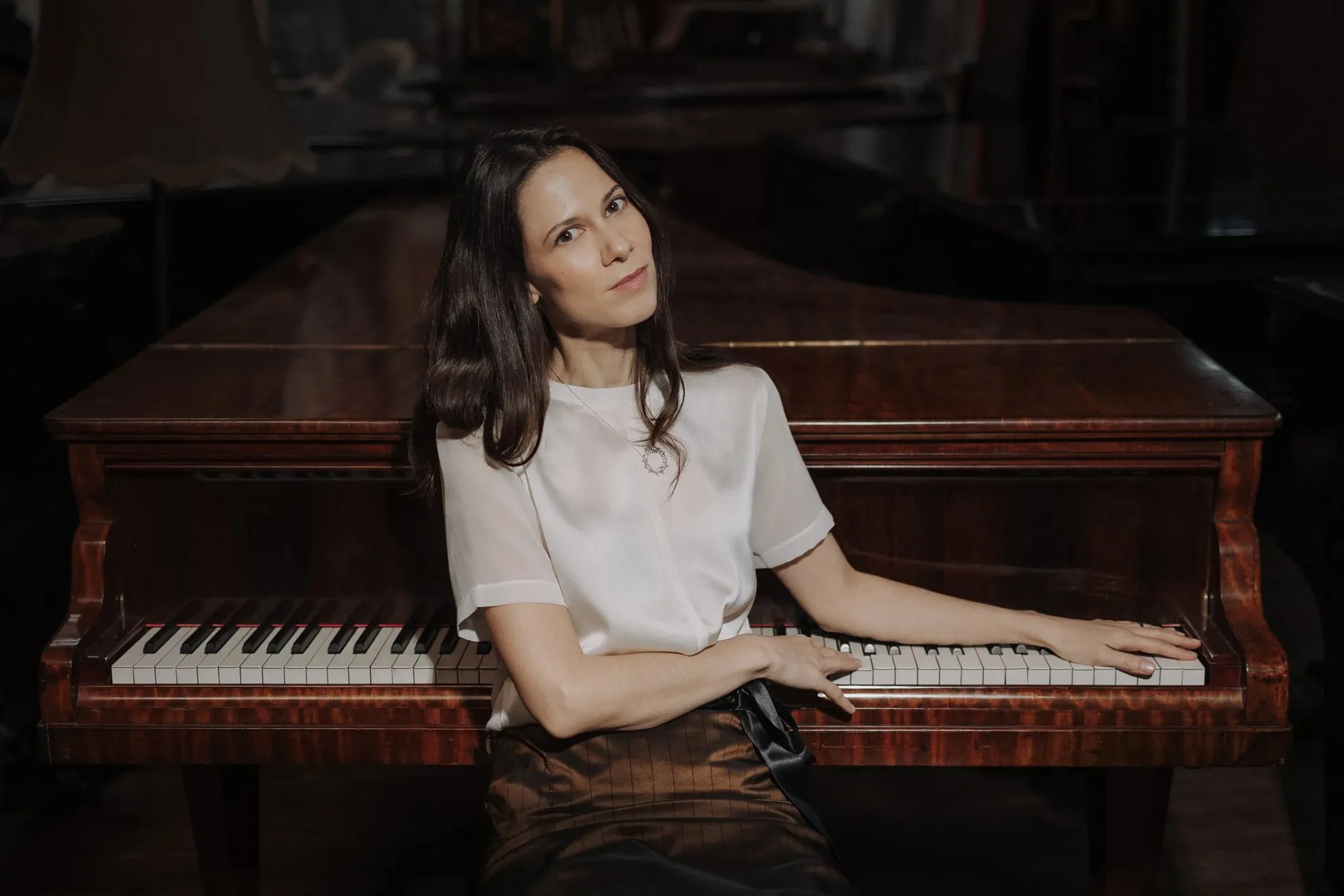 Woman seated at a polished wooden grand piano, hands on the keys in a dimly lit room.