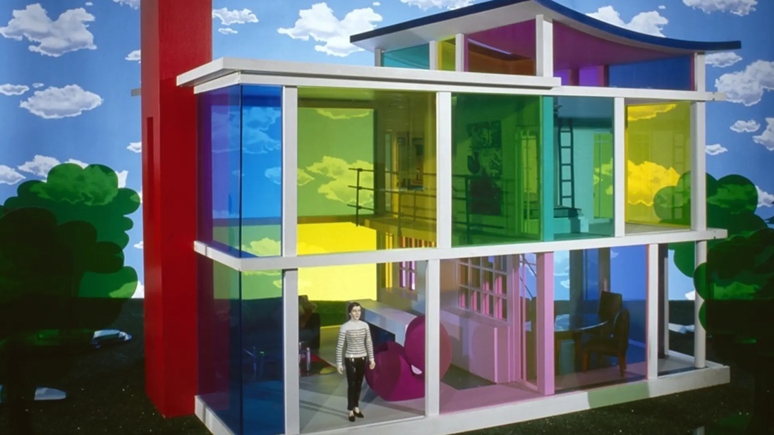 Colorful glass-walled house sculpture with rainbow-tinted panels and a person standing outside.