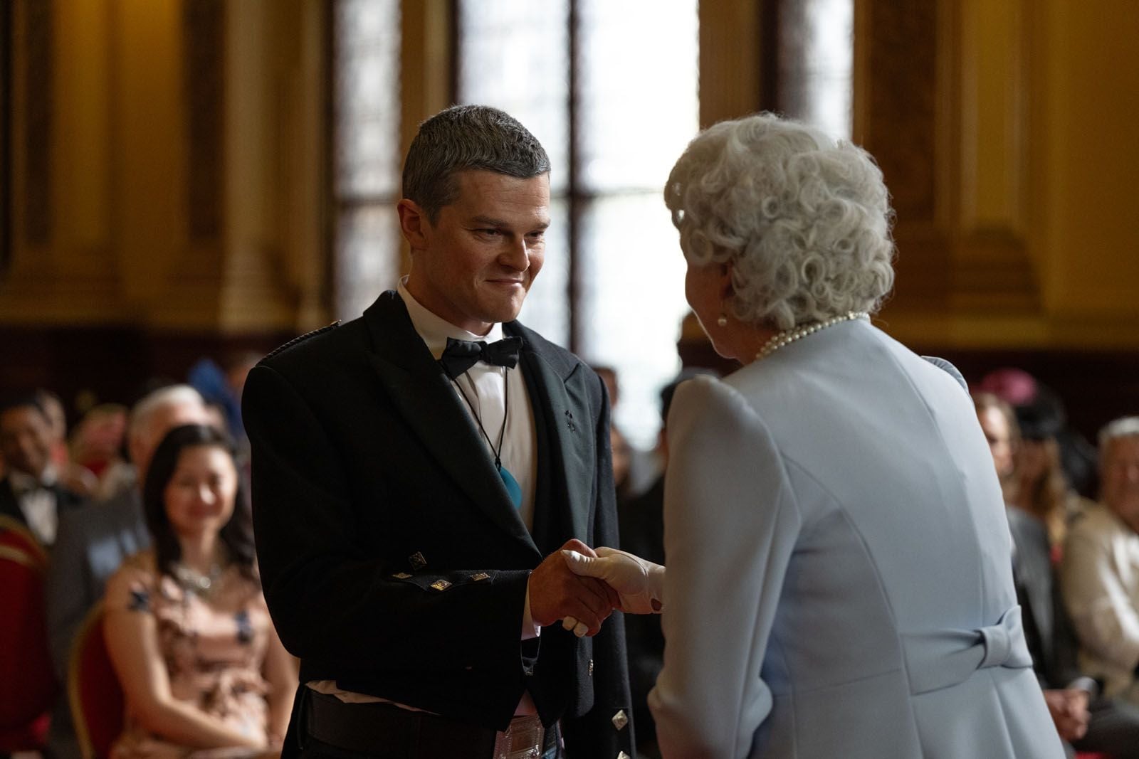 A man in formal military dress shakes hands with an older woman in a light blue suit at a ceremony, with seated guests watching in the background.