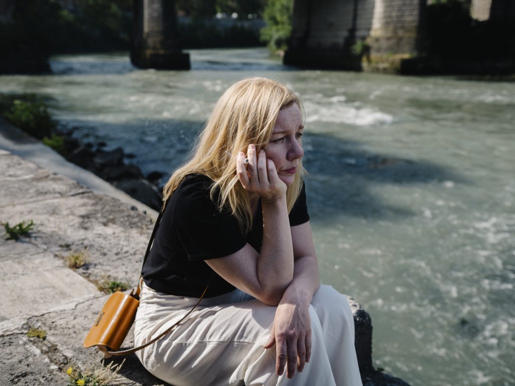 BACHMANN (Regina Schilling, zeroone film GmbH) - IN München - Das Stadtmagazin Woman sitting by a river, smoking a cigarette, wearing a black top and beige skirt, with an orange bag beside her by the rocky shore.