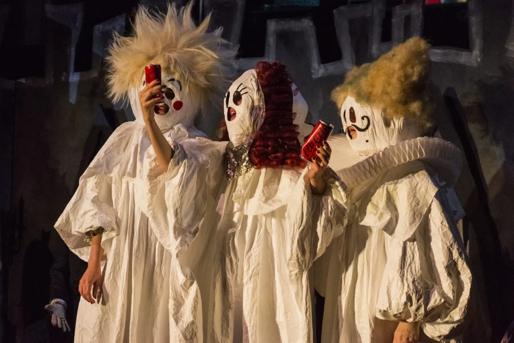 13_I WANNA CONTACT THE LIVING_Liv Stapelfeldt, Marlene Markt, Maximiliane Haß (c) Jakob Schnetz - IN München - Das Stadtmagazin Three performers in white sheet costumes with clown masks on a dark stage, each holding a red Coca‑Cola can.