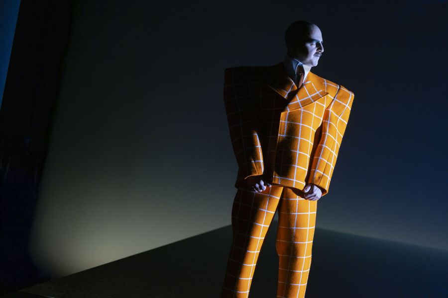 16_Pioniere_in_Ingolstadt_MVT_Pascal Fligg(c)MarcellaRuizCruz - IN München - Das Stadtmagazin Mannequin wearing a bold orange checkered suit, standing on a dark stage with dramatic side lighting.