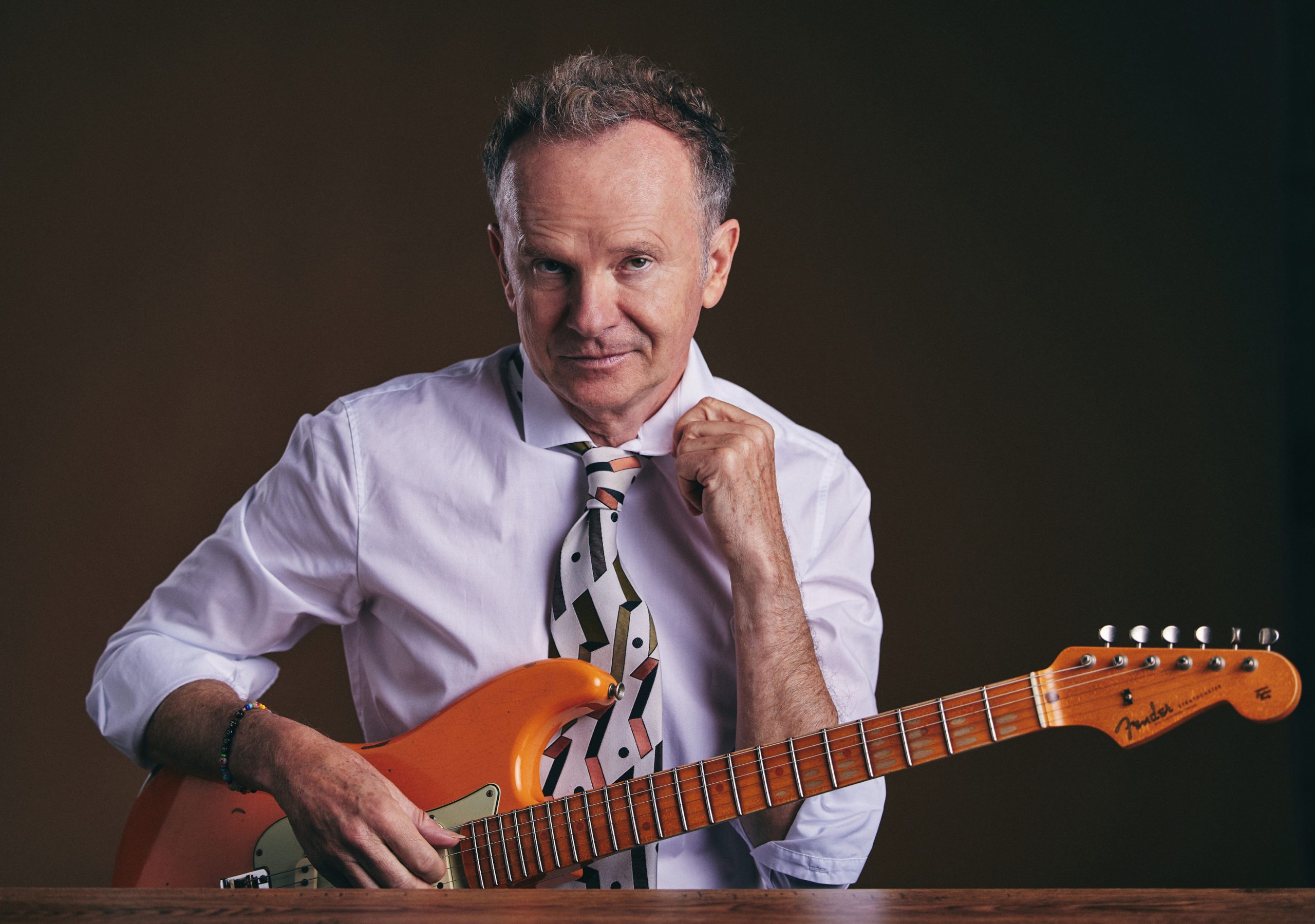 Man in a white shirt and patterned tie playing an orange Fender Stratocaster, resting hands on the guitar neck indoors.