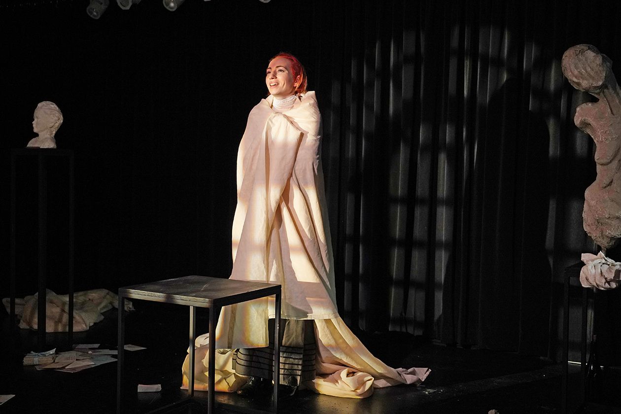 5220_sth3_freiheit_stueckbild_06 - IN München - Das Stadtmagazin Actor on a dim stage wearing a pale pink cape, smiling under warm lights with bust sculptures nearby and papers on the floor.