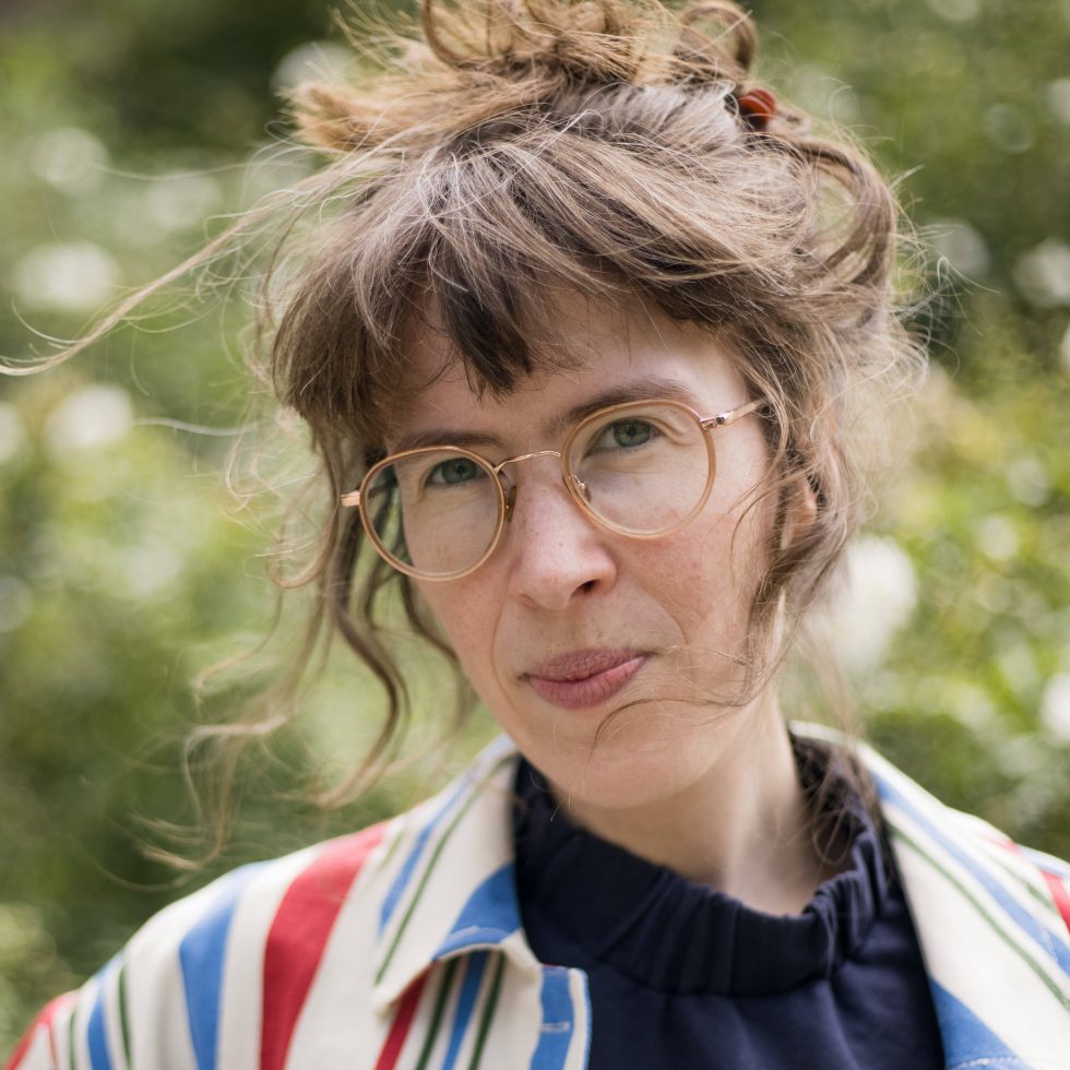 Bach_Kathrin_cJulia_Vogel_klein-kl-980x980 - IN München - Das Stadtmagazin Woman with round glasses in a sunny outdoor garden, hair tied up in a casual bun, wearing a striped shirt over a dark top.