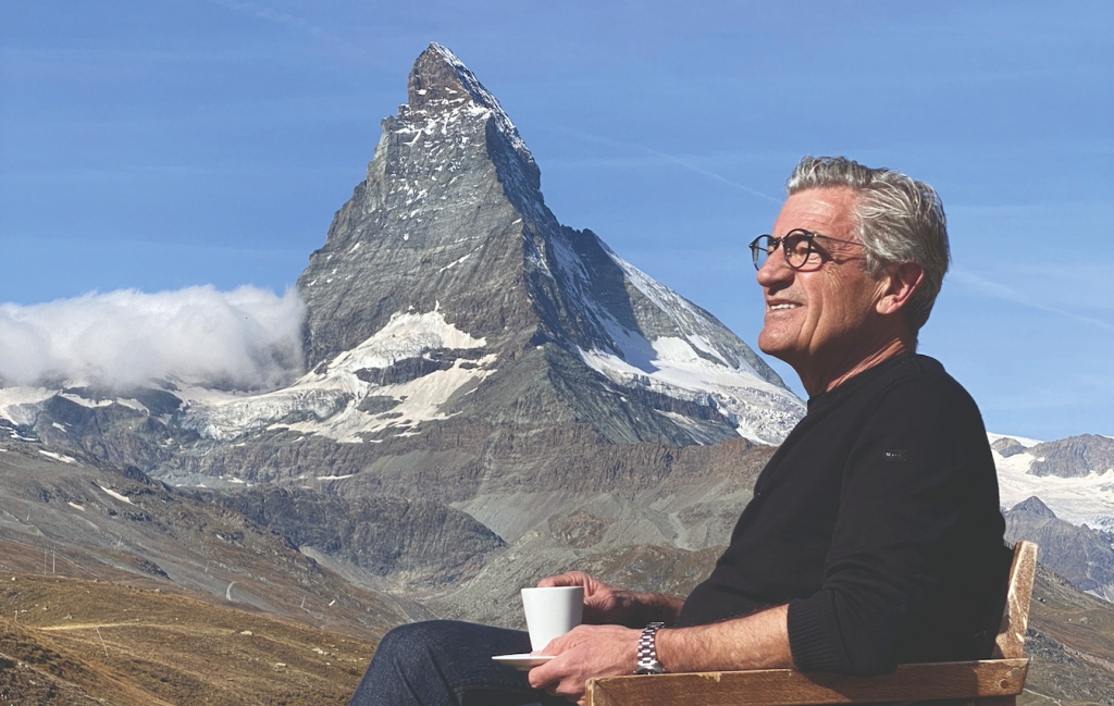 Older man with glasses sitting on a wooden bench, holding a white cup, with a snow-capped mountain in blue sky behind him.