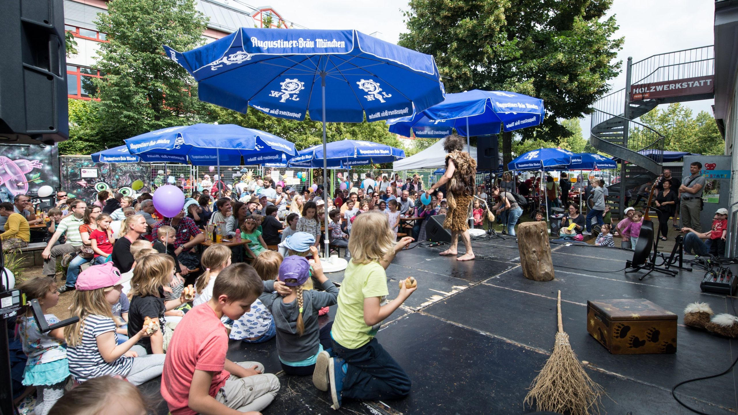 Outdoor festival scene with children watching a performer in a fur costume on a small stage under blue umbrellas.