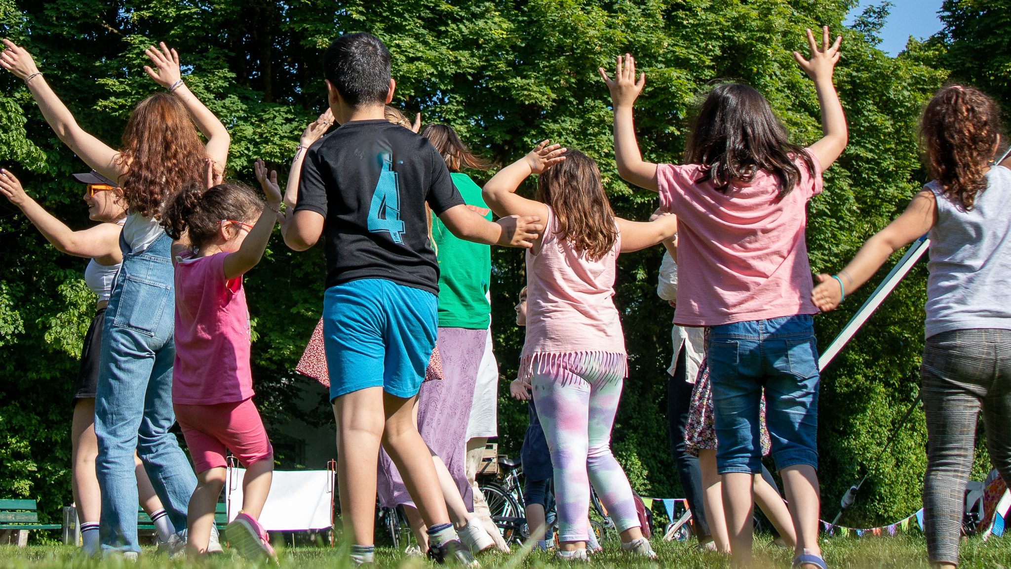 Ferienkurs-–-Tanzreise-Zeit - IN München - Das Stadtmagazin Group of children outdoors with arms raised, dancing/celebrating in a sunny park.