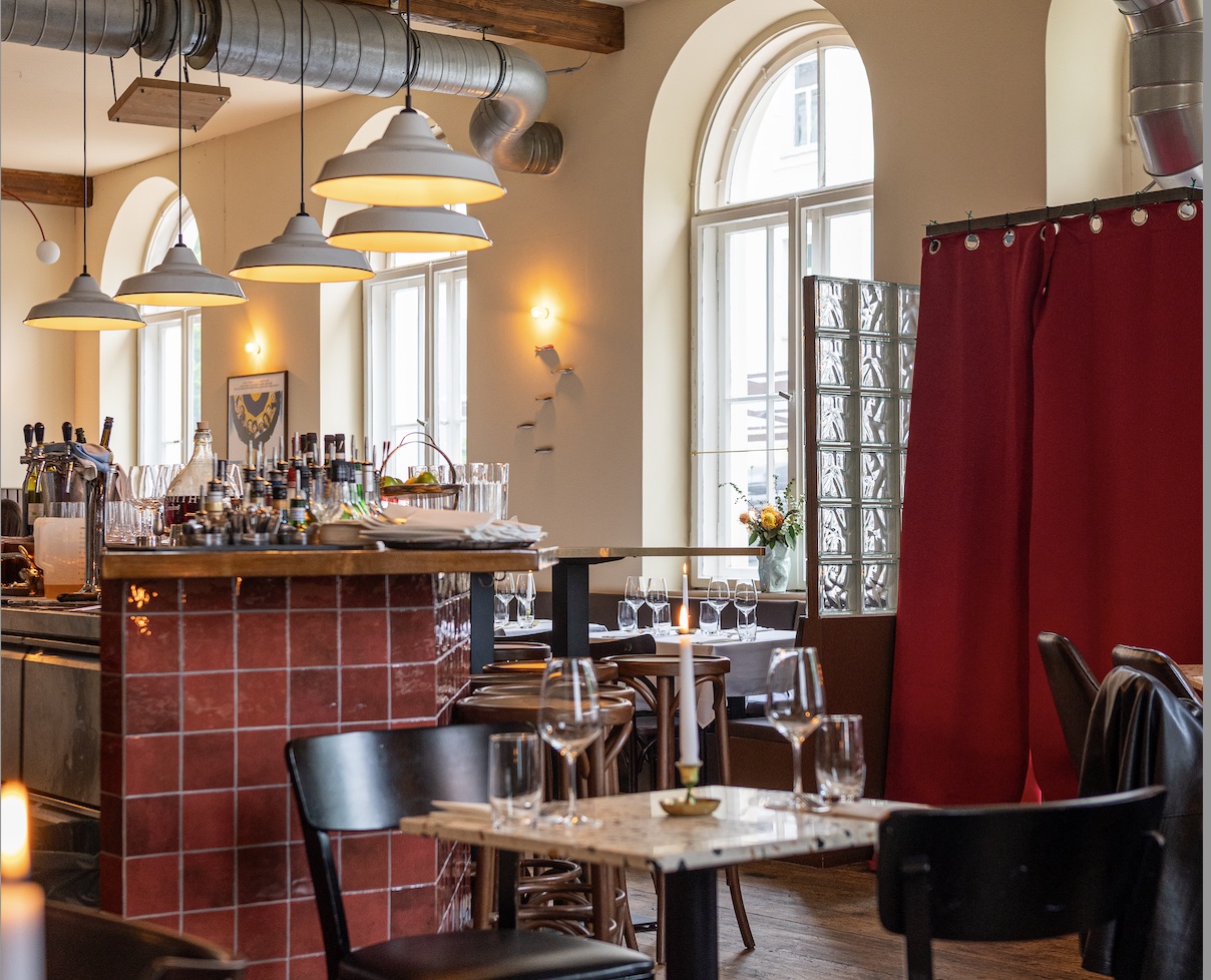 Cozy restaurant interior with a tiled red bar, wooden chairs, hanging pendant lights, large arched window, and candlelit tables.