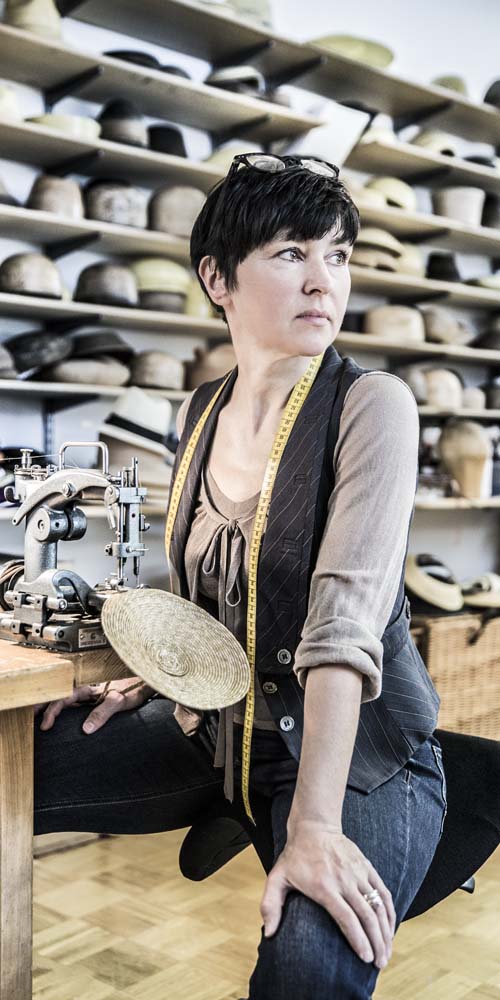 Woman sewing a straw hat at a sewing machine in a hat shop, shelves full of hats in the background.