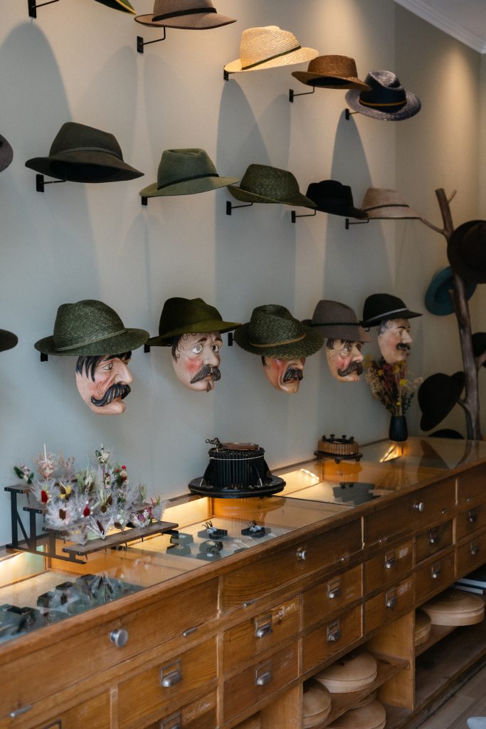 Wall display of felt and straw hats mounted on a pale wall, with decorative masks hanging beneath them and a wooden counter below with small accessories.