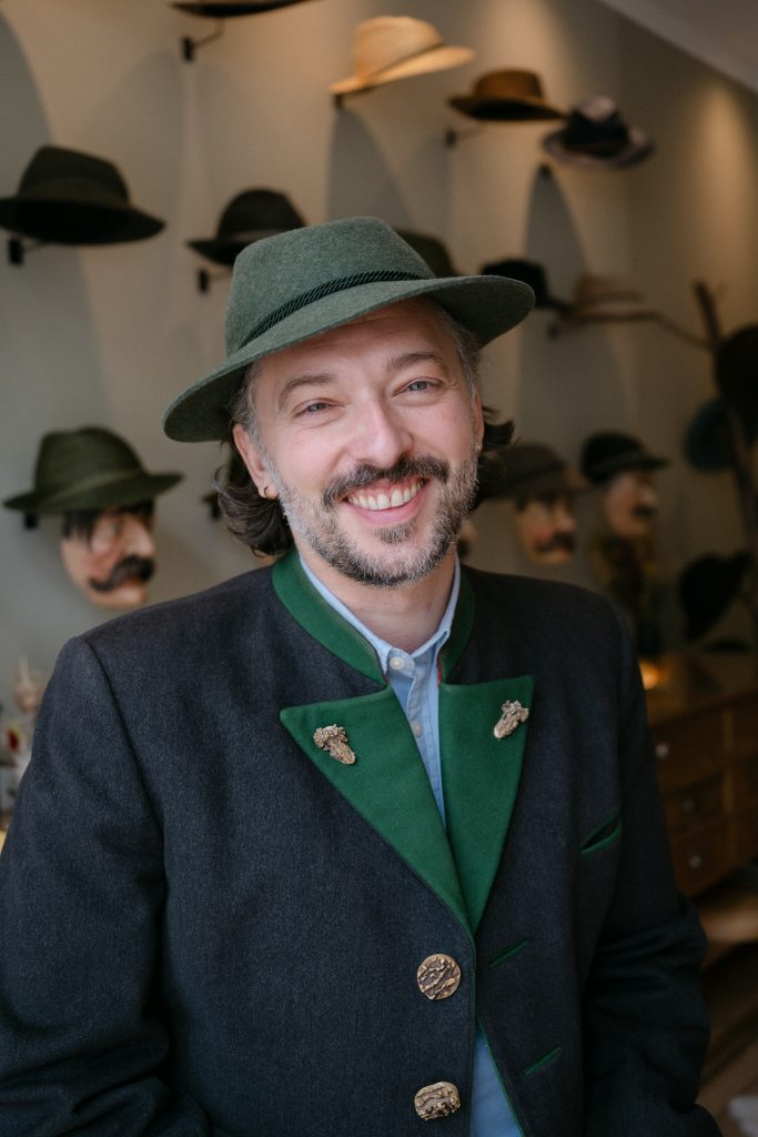 Smiling man wearing a green hat and dark blazer with green lapels, decorated with pins; hat display wall in background.
