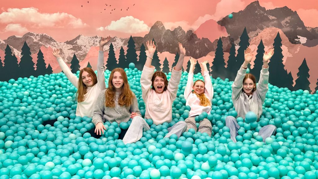 Five children sit in a turquoise ball pit, arms raised, against a pink mountain mural backdrop with trees and mountains behind them.