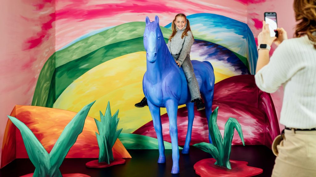 Girl smiling rides a blue horse statue in a vivid rainbow-painted exhibit; a person in foreground takes a photo with a phone.