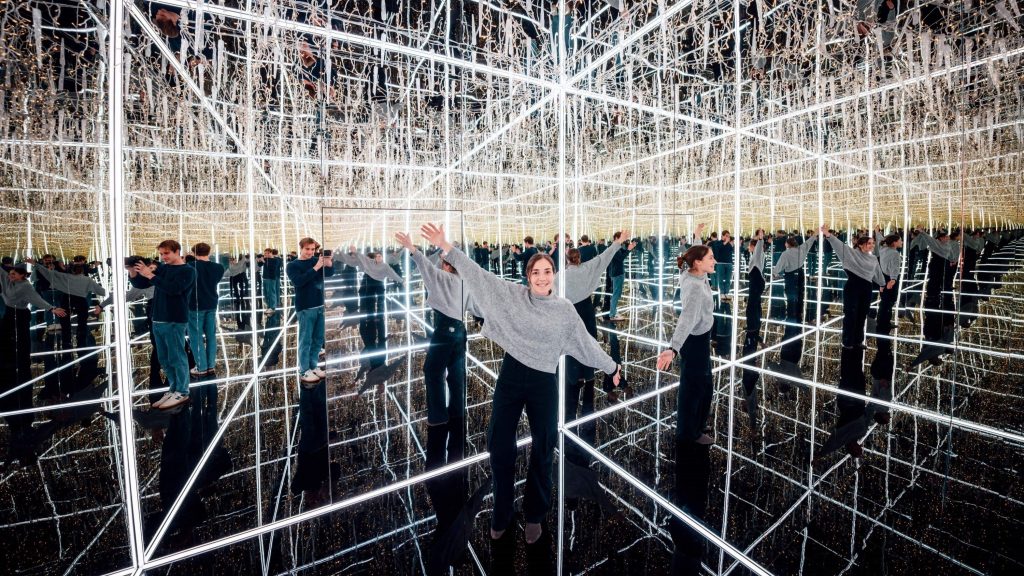 People posing inside a mirrored room filled with crisscrossing LED light beams, creating endless reflections.