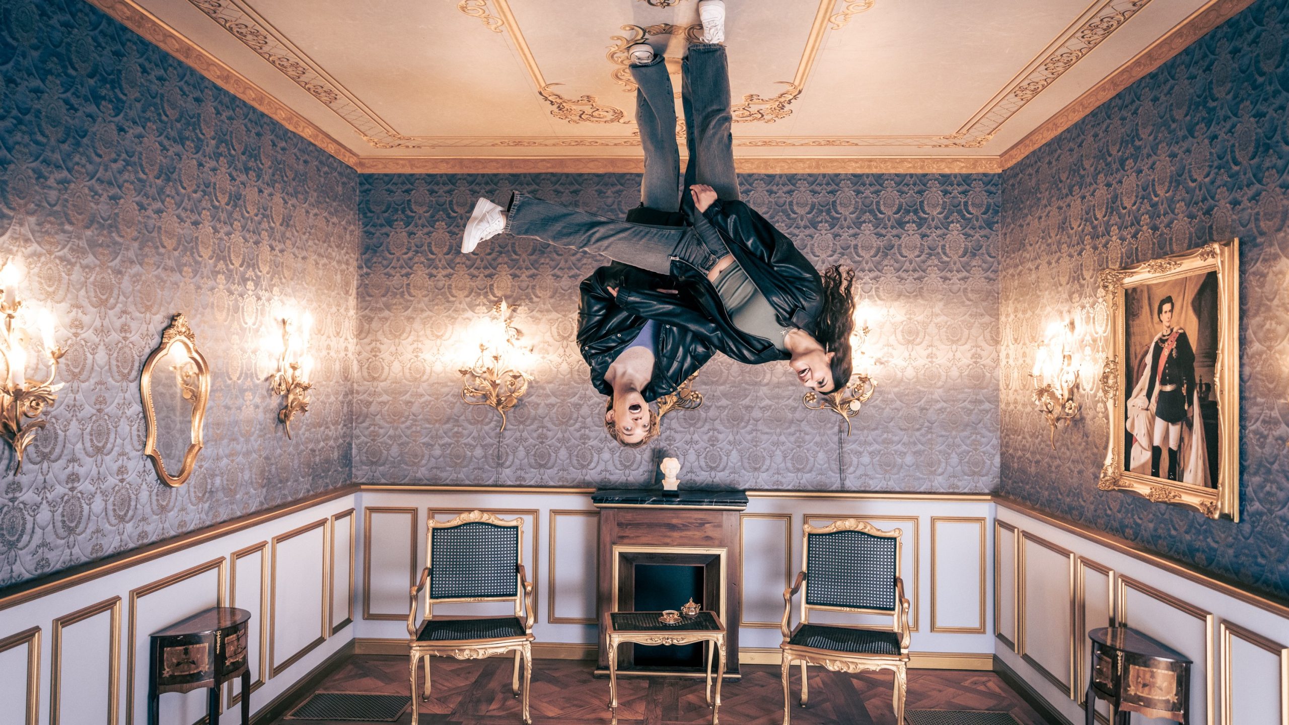 Upside-down ornate room with two people hanging from the ceiling in leather jackets amid blue damask wallpaper and gilded trim. Portrait on the wall and gilded chairs surround the scene.