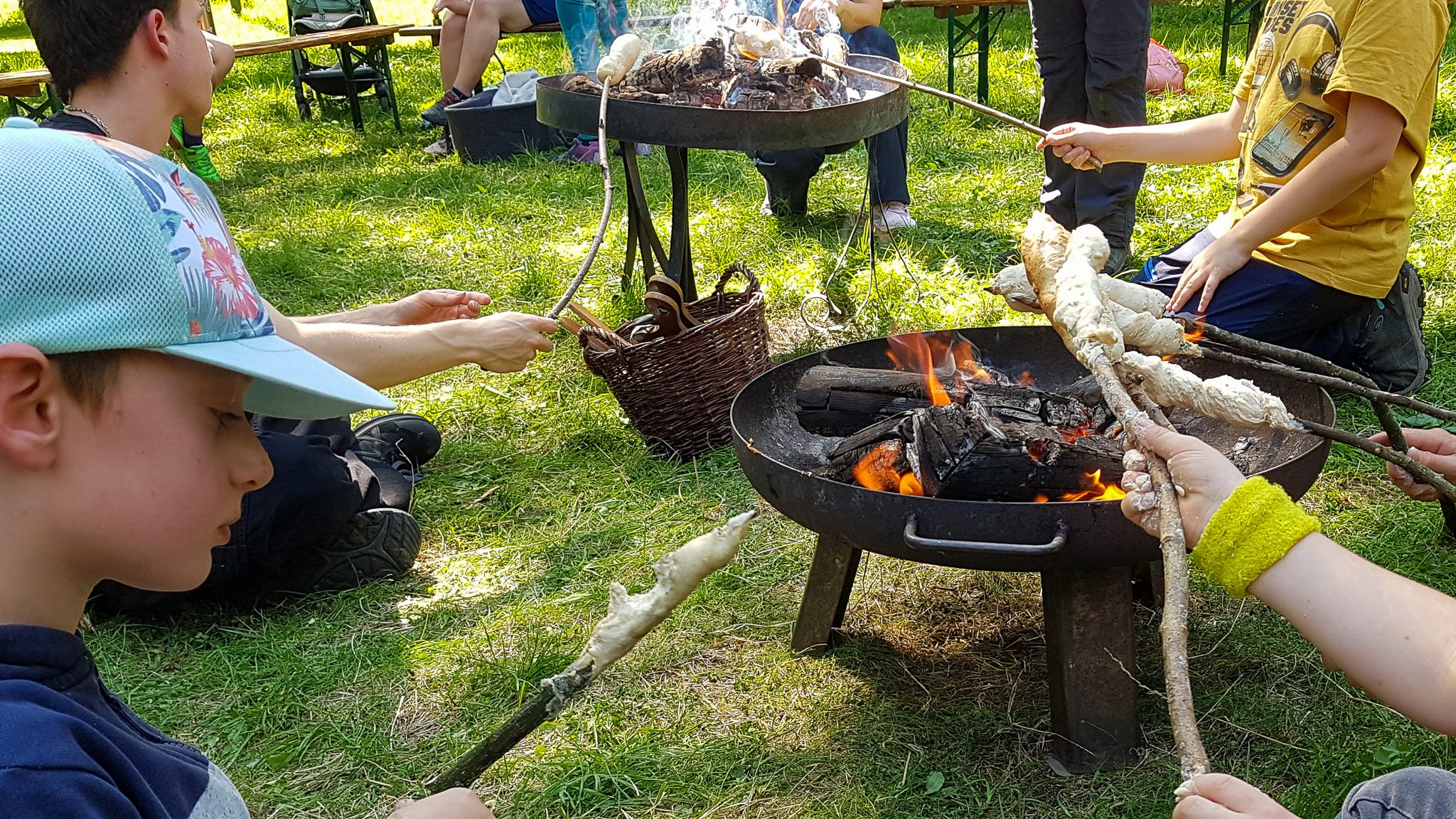 Mammut-Stein-Feuerschein-–-Jagen-und-Sammeln - IN München - Das Stadtmagazin Children sitting on the grass around a campfire, roasting marshmallows on sticks in a sunny park.