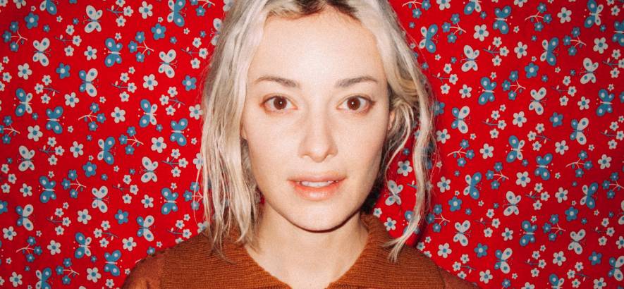 Portrait of a woman with shoulder-length blonde hair wearing an orange/brown sweater, standing against a bright red floral-patterned background.