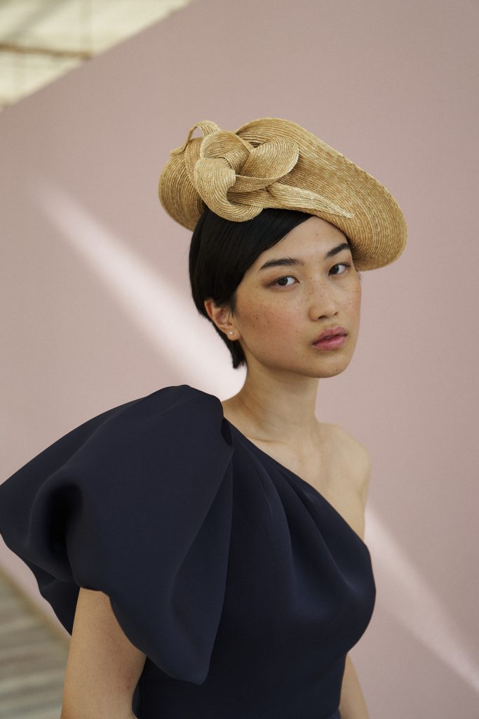 Portrait of a woman wearing a woven straw hat with decorative loops, against a muted pink backdrop.