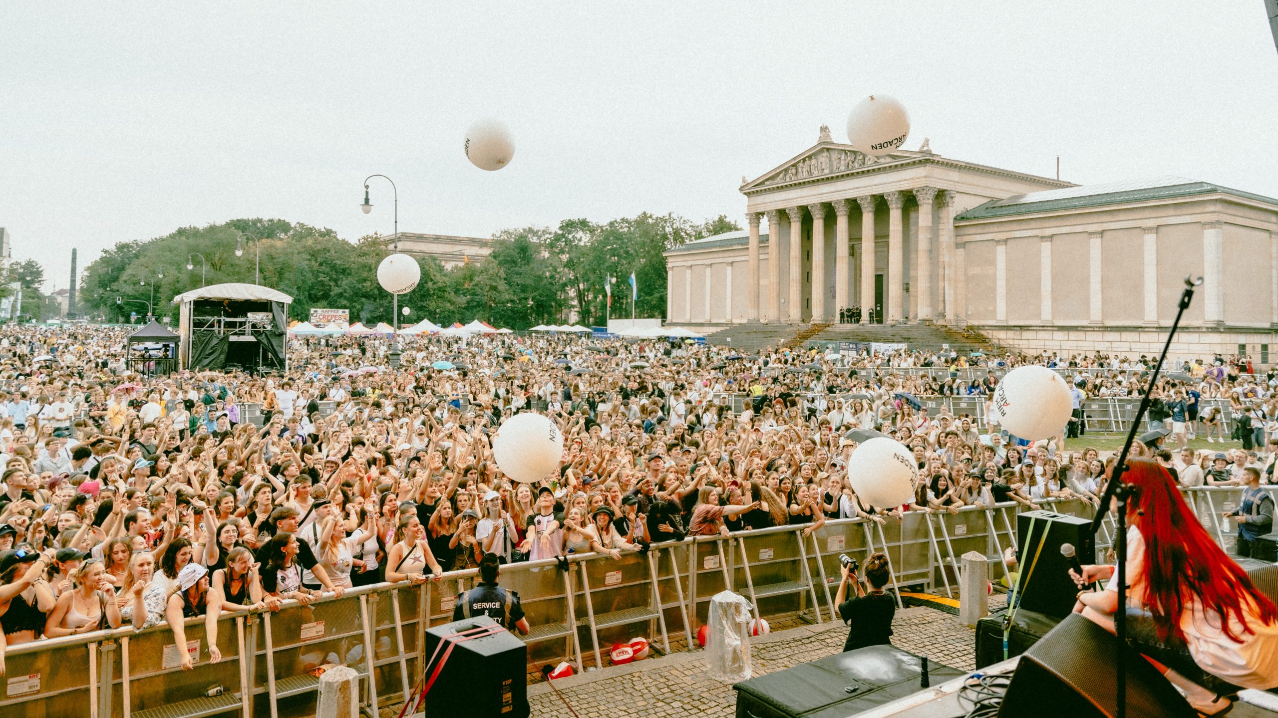 Massive outdoor concert crowd with hands raised in front of a neoclassical building with tall columns and balloons in the sky, stage visible on the right.