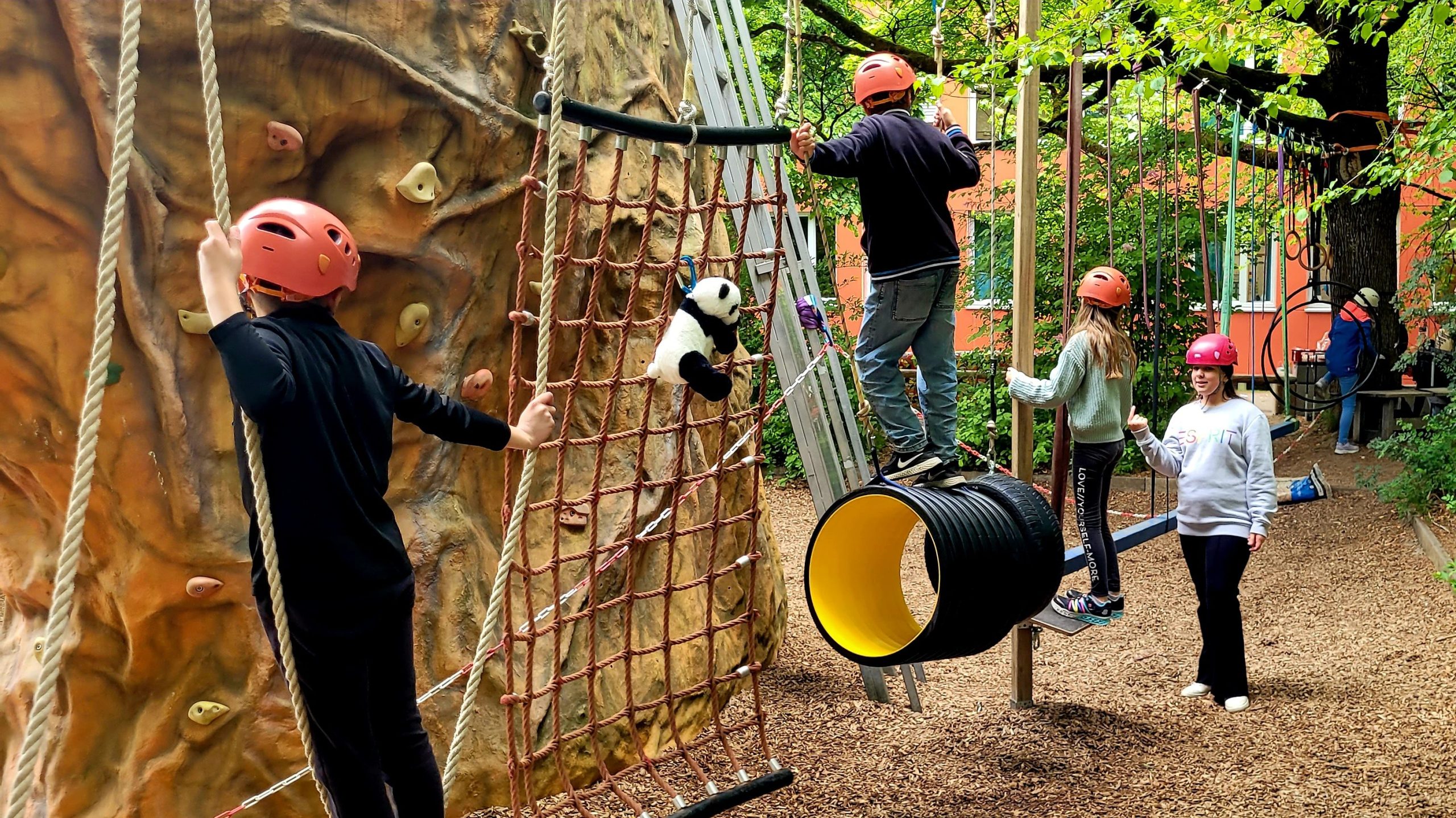 Children in helmets navigate a rope climbing wall and obstacle course outdoors, with a panda plush toy hanging from the net.