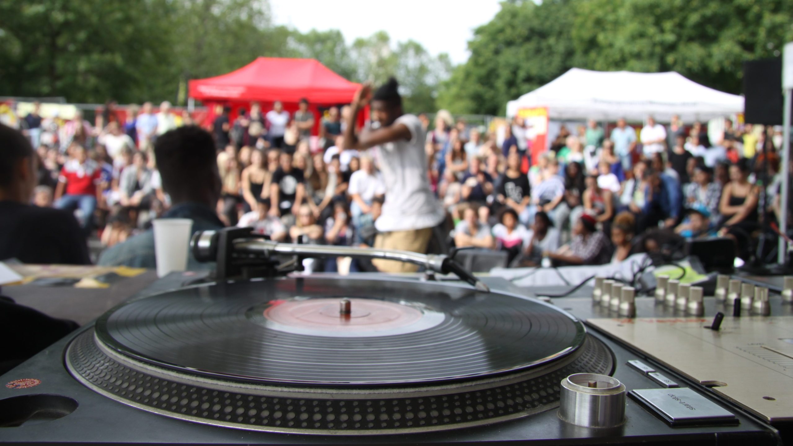 DJ performing on a turntable at an outdoor festival with a large crowd in the background, tents visible behind the audience