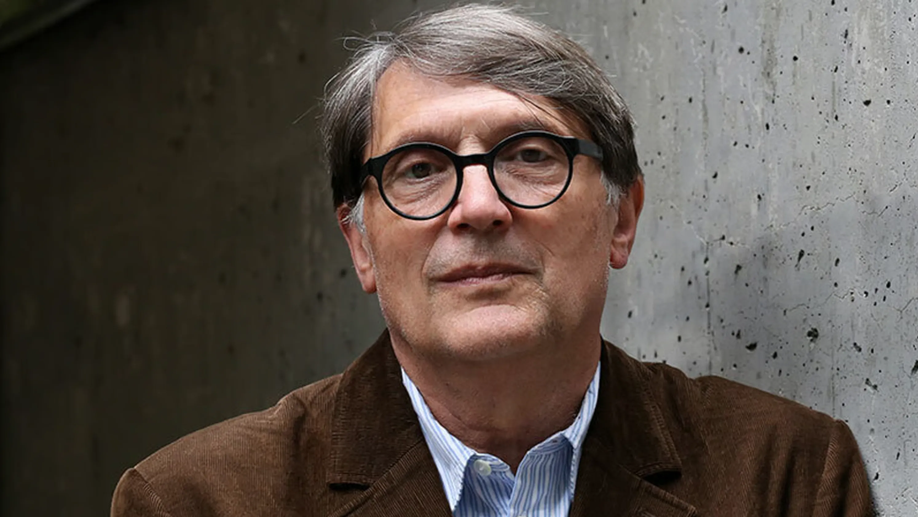 Screenshot 2026-06-15 111804 - IN München - Das Stadtmagazin Portrait of an older man with gray hair and black round glasses, wearing a brown jacket, against a concrete wall background.