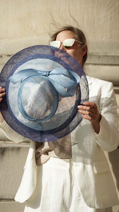 Person in a white suit holds a large, blue wide-brimmed hat in front of their face with reflective sunglasses nearby.