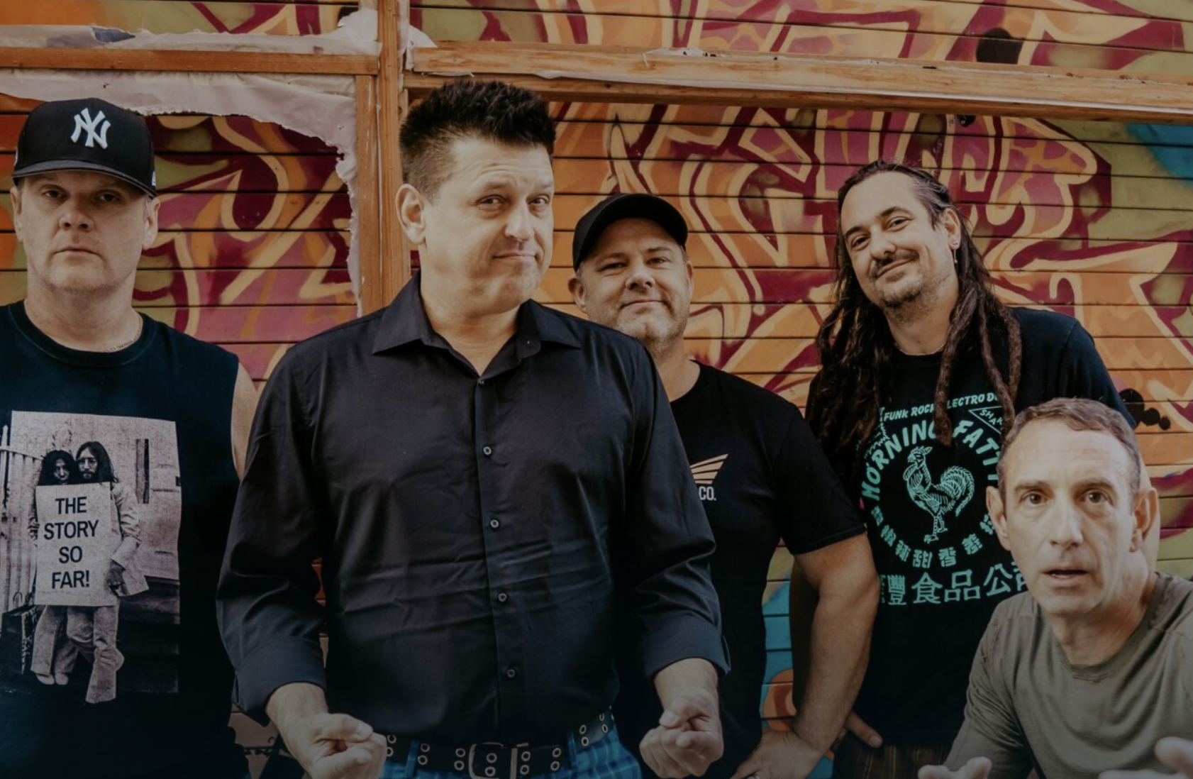Five men pose in front of a colorful graffiti wall, wearing casual clothes and looking at the camera.