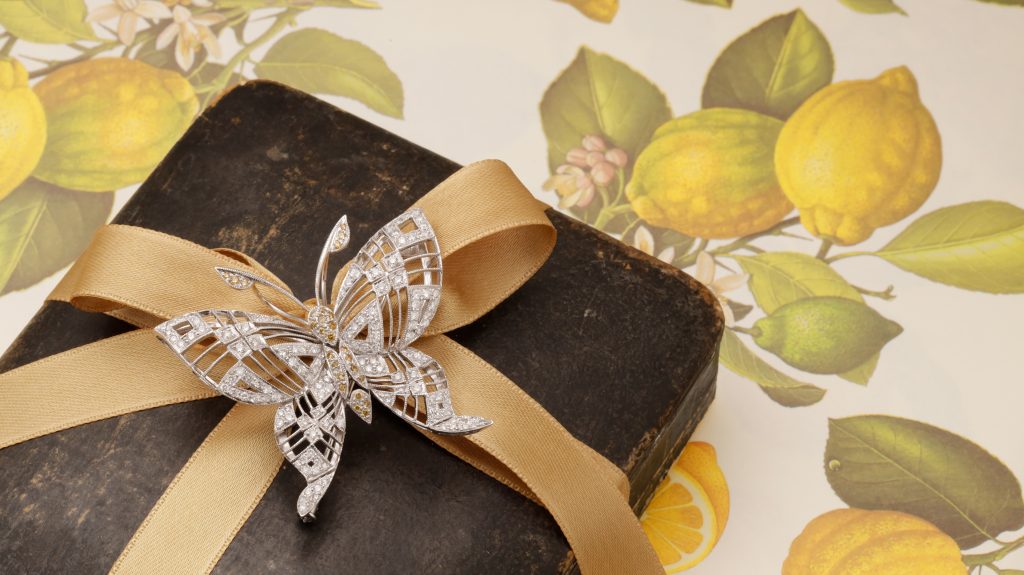 Gift box wrapped with a gold ribbon and a silver jeweled butterfly bow, set against a yellow citrus-themed backdrop.