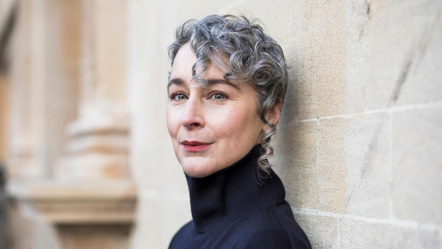 Portrait of a woman with short gray curly hair wearing a black turtleneck, leaning against a stone wall.