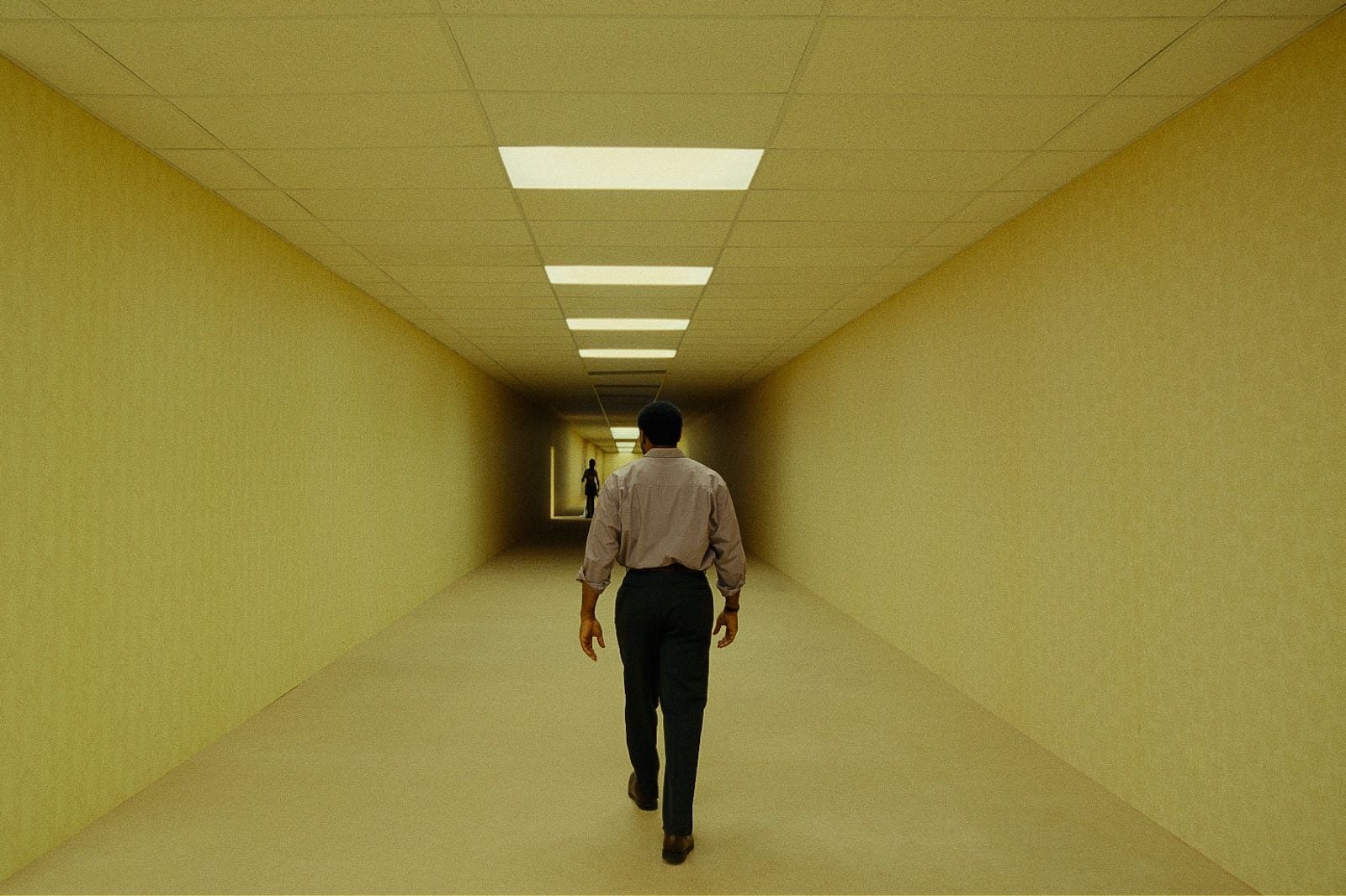Man in a light shirt walks away down a long, empty beige hallway toward a distant figure at the end.