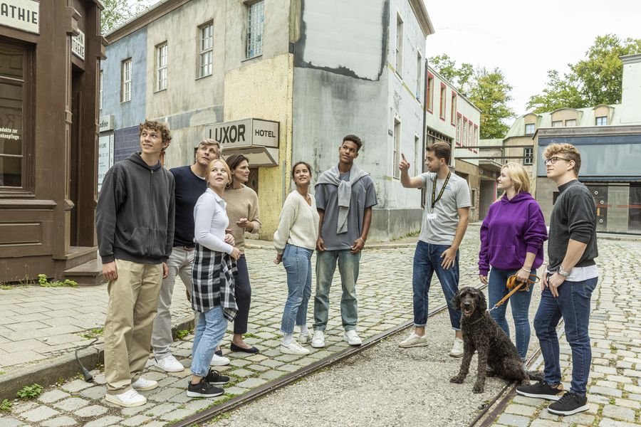 dog days programm muenchen - IN München - Das Stadtmagazin Group of diverse young adults standing on a cobblestone street with a dog on a leash, looking toward a speaker off to the right near old buildings.